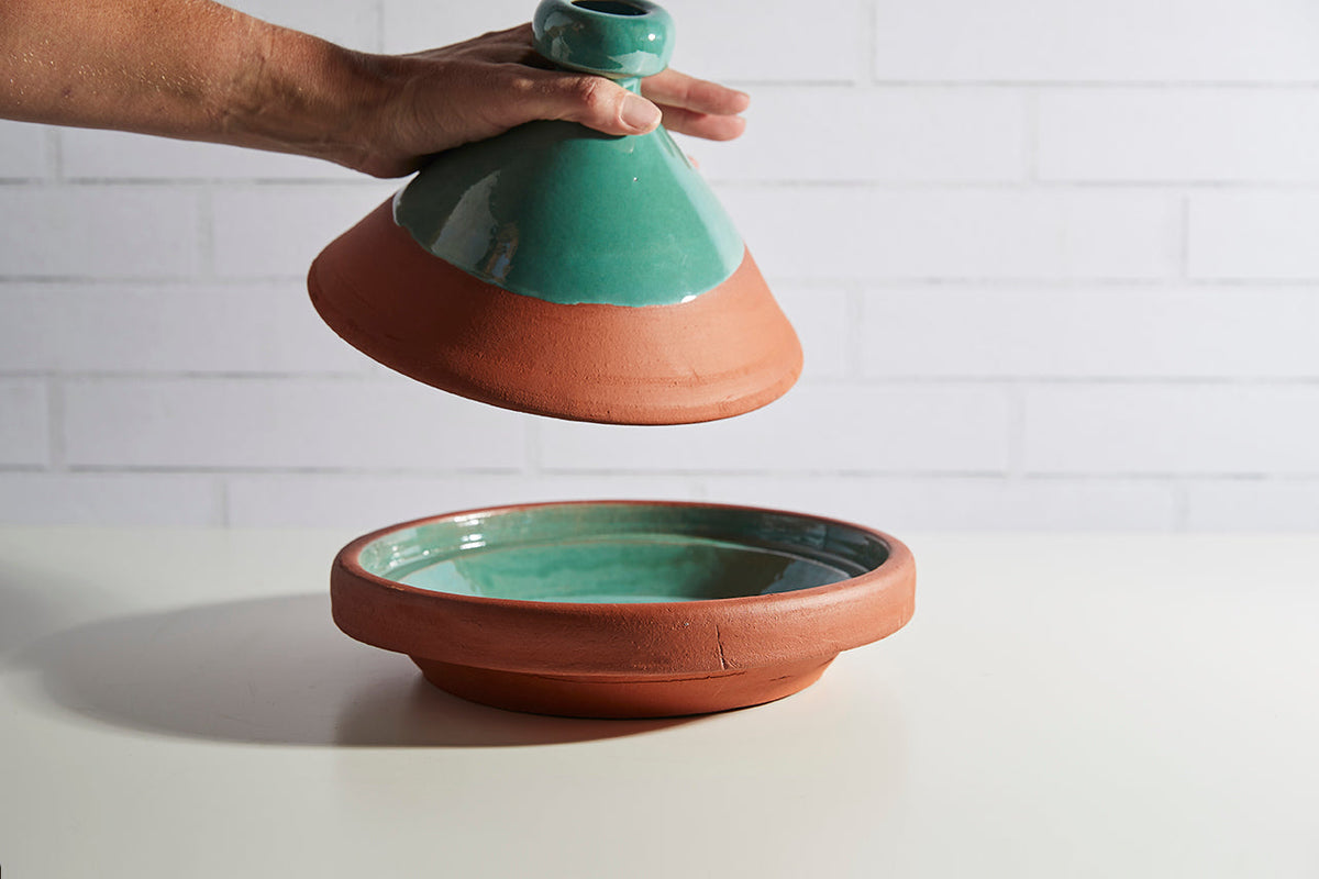 Two-piece tagine with terracotta base and turquoise cone-shaped lid being held by hand.