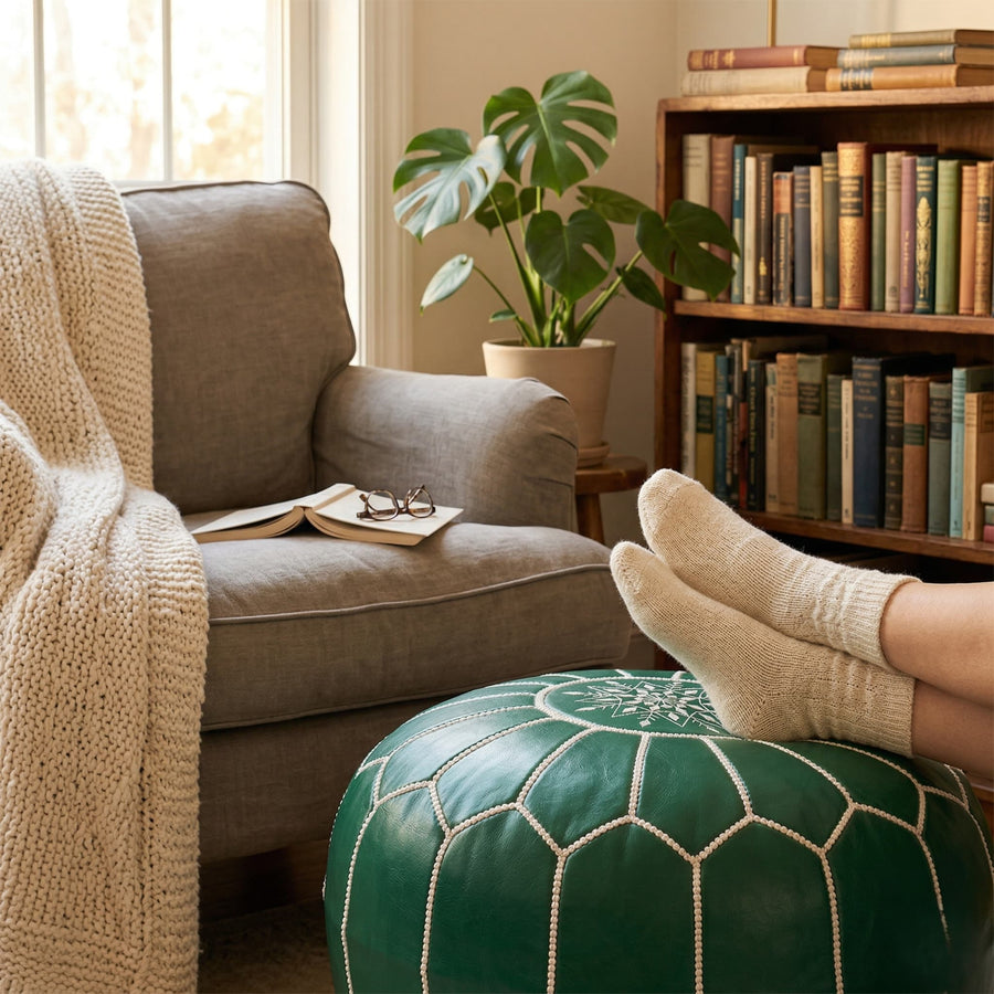 Feet in wool socks resting on the green Moroccan pouf in a sunlit reading corner with a linen armchair and bookshelves
