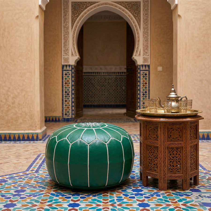 Green leather round pouf on zellige tilework in a Moroccan riad courtyard next to a carved wooden table with a brass tea set