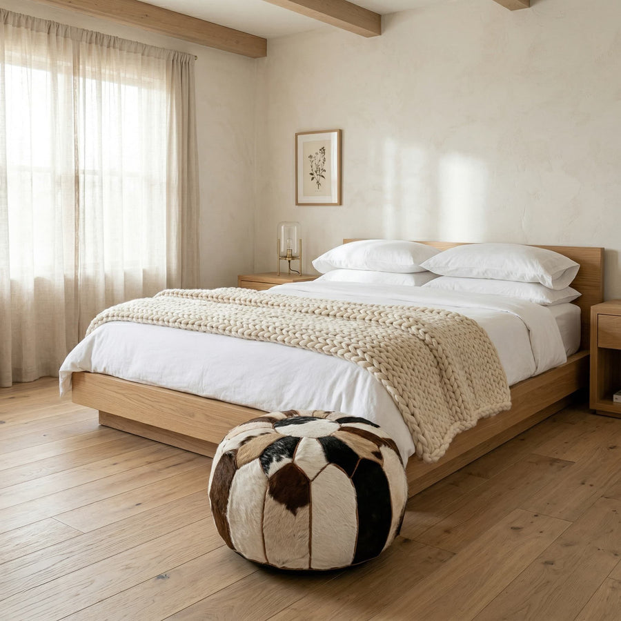 Cowhide round pouf used as a footstool at the foot of an oak bed in a bright minimal bedroom