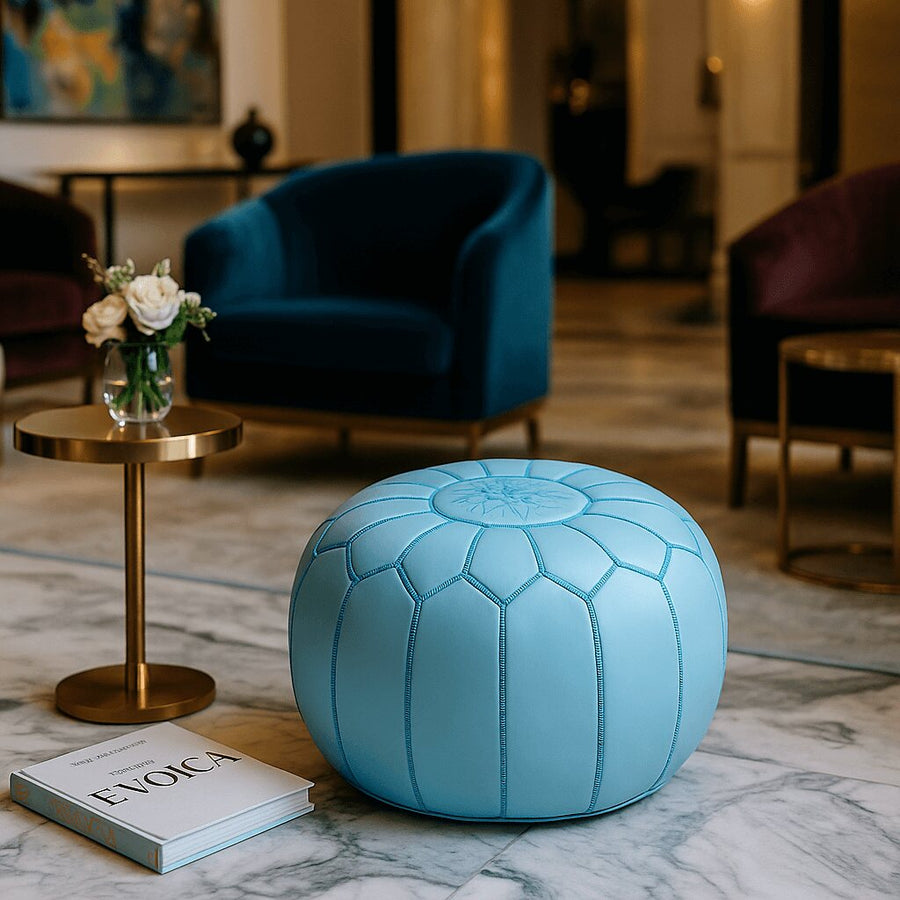 Sky blue leather round pouf on marble flooring in a hotel lounge with navy velvet armchairs and a gold side table