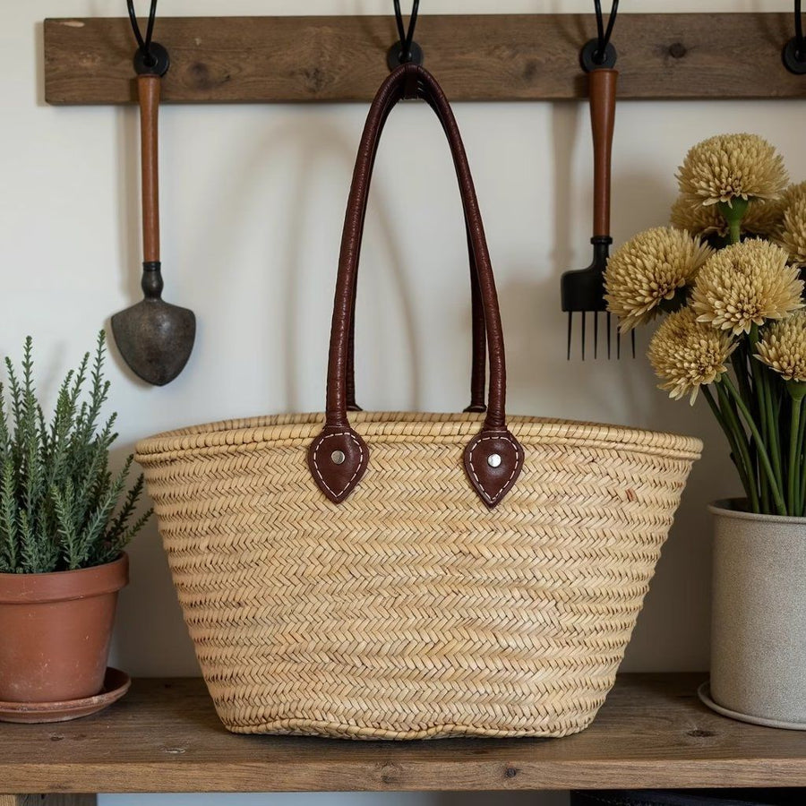Palm leaf basket tote hanging by its long leather shoulder handles from a wooden peg rail