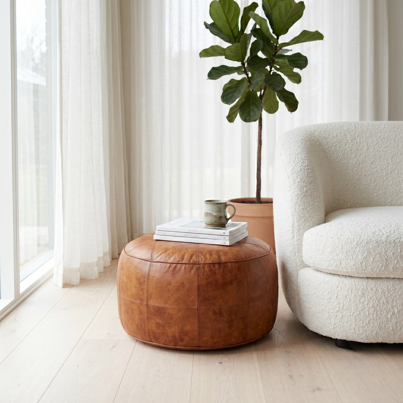 Tan leather floor ottoman beside a boucle armchair and fiddle leaf fig in a minimalist living room